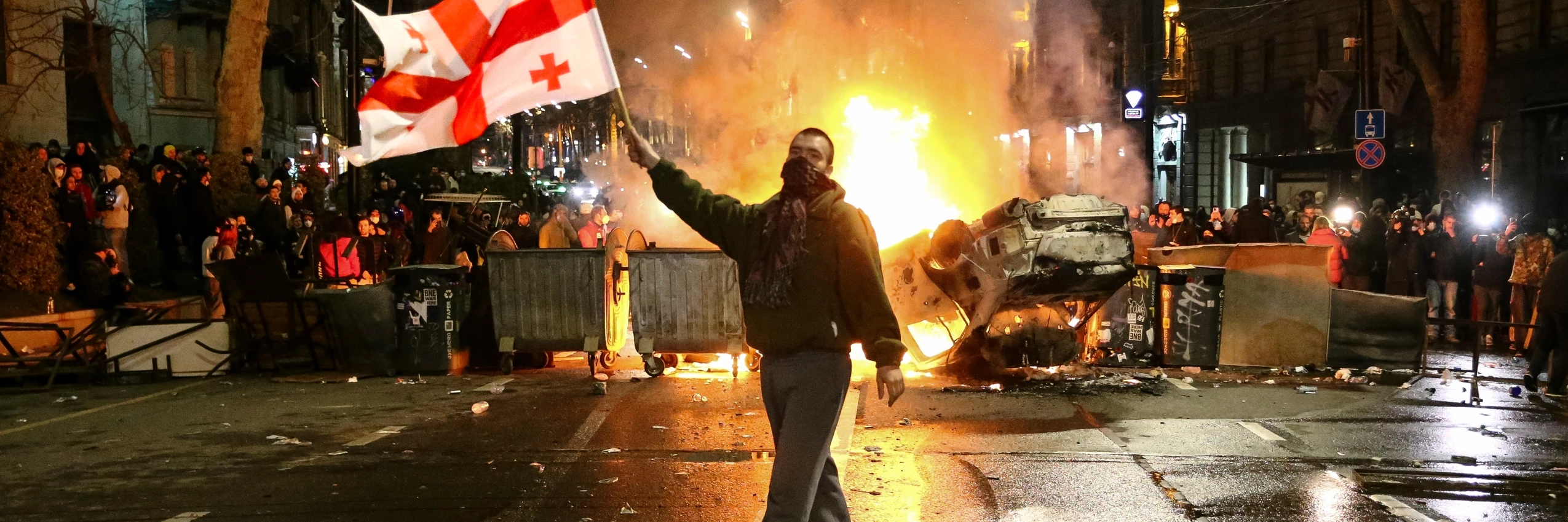 Ein Mann schwenkt eine georgische Nationalflagge vor einer brennenden Barrikade während andere unweit des georgischen Parlamentsgebäudes in Tbilisi, Georgien, protestieren. © Zurab Tsertsvadze / AP / Picturedesk.Com