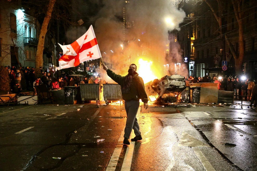 Ein Mann schwenkt eine georgische Nationalflagge vor einer brennenden Barrikade während andere unweit des georgischen Parlamentsgebäudes in Tbilisi, Georgien, protestieren. © Zurab Tsertsvadze / AP / Picturedesk.Com