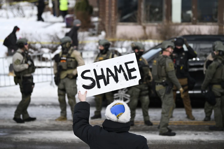 Eine Person hält ein Schild mit der Aufschrift "Shame" (Schande) hoch, während Bundespolizisten nach einer mutmaßlichen Schießerei durch einen ICE-Beamten am 7. Januar 2026 in Minneapolis, Minnesota, am Tatort arbeiten @ Stephen Maturen/Getty Images