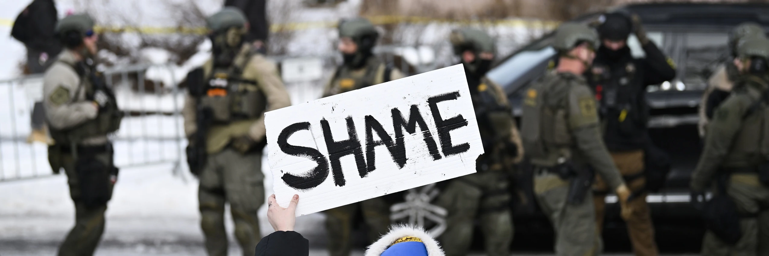 Eine Person hält ein Schild mit der Aufschrift "Shame" (Schande) hoch, während Bundespolizisten nach einer mutmaßlichen Schießerei durch einen ICE-Beamten am 7. Januar 2026 in Minneapolis, Minnesota, am Tatort arbeiten © Stephen Maturen/Getty Images