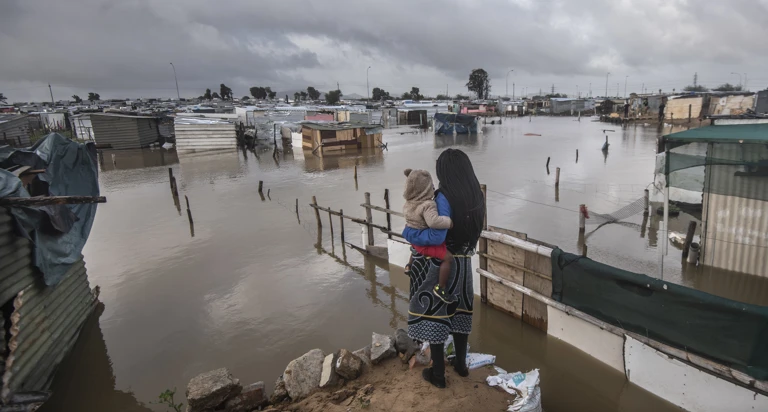 Eine Frau steht mit ihrem Baby im Arm vor einer überfluteten informellen Siedlung in Kapstadt, Südafrika (2021). @ Brenton Geach/Gallo Images (PTY) LTD 