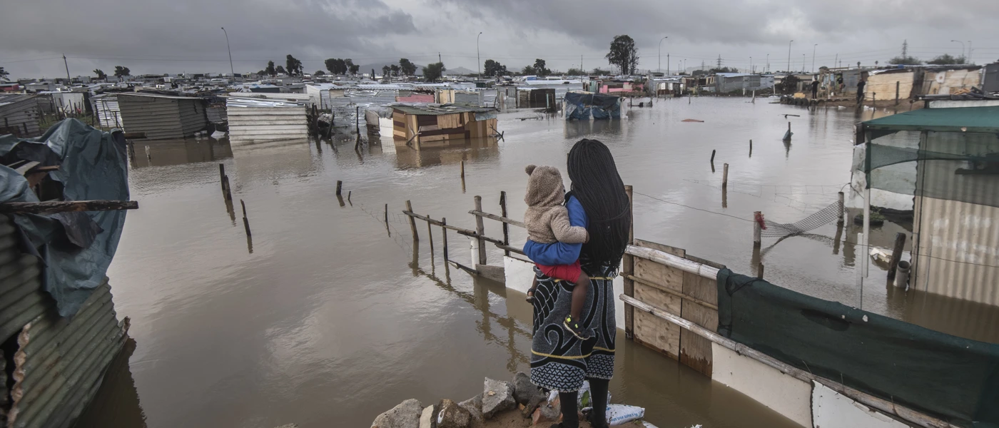 Eine Frau steht mit ihrem Baby im Arm vor einer überfluteten informellen Siedlung in Kapstadt, Südafrika (2021). © Brenton Geach/Gallo Images (PTY) LTD