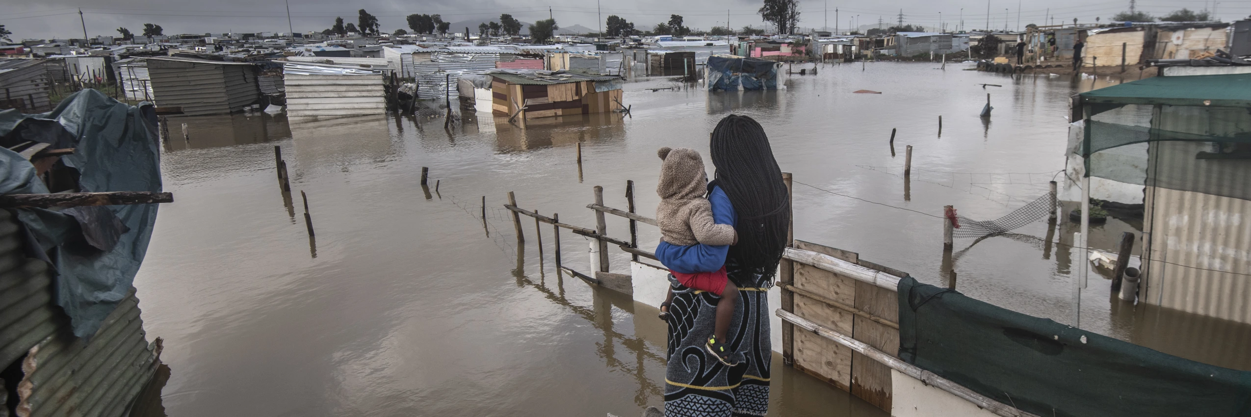 Eine Frau steht mit ihrem Baby im Arm vor einer überfluteten informellen Siedlung in Kapstadt, Südafrika (2021). © Brenton Geach/Gallo Images (PTY) LTD