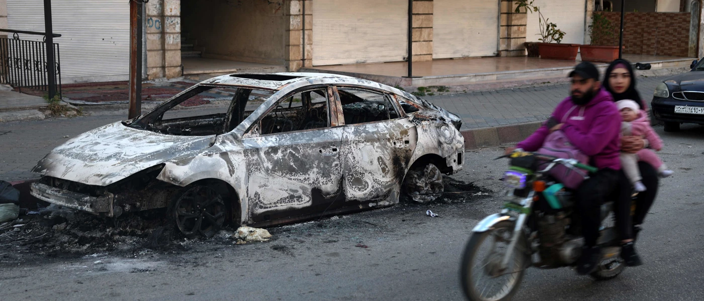 12. März 2025: Eine Familie fährt an einem verkohlten Auto in der Stadt Jableh in der syrischen Küstenprovinz Latakia vorbei. © Omar Haj Kadour, AFP, picturedesk.com