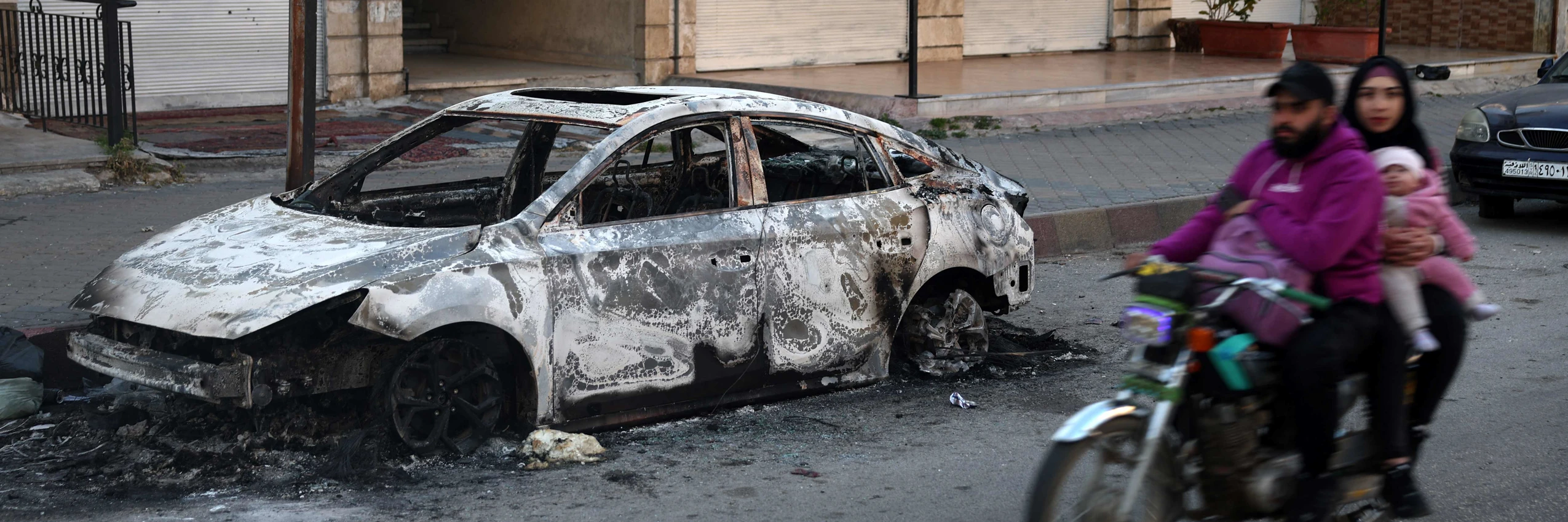 12. März 2025: Eine Familie fährt an einem verkohlten Auto in der Stadt Jableh in der syrischen Küstenprovinz Latakia vorbei. © Omar Haj Kadour, AFP, picturedesk.com