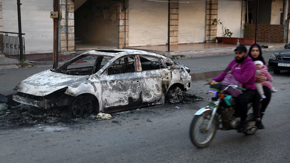 12. März 2025: Eine Familie fährt an einem verkohlten Auto in der Stadt Jableh in der syrischen Küstenprovinz Latakia vorbei. © Omar Haj Kadour, AFP, picturedesk.com
