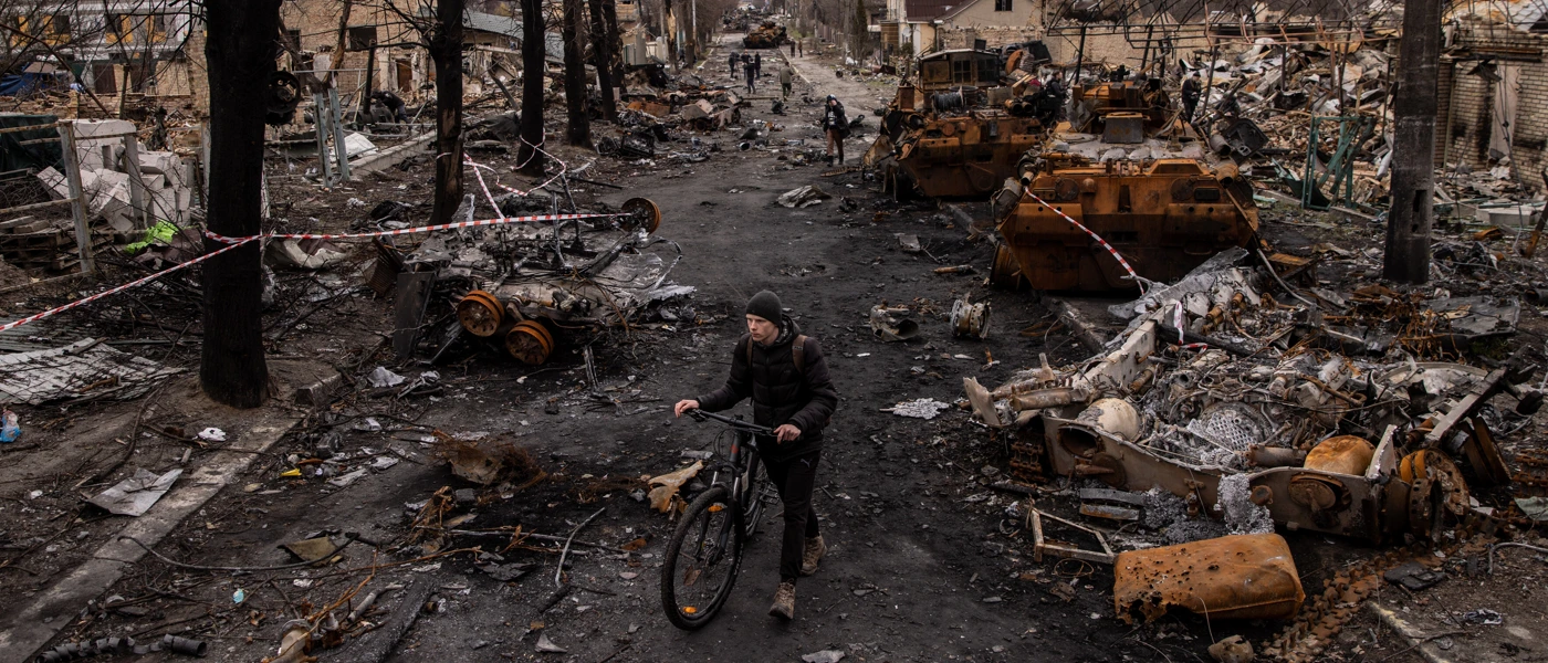 Ein Mann schiebt sein Fahrrad durch Trümmer und zerstörte Militärfahrzeuge auf einer Straße in Bucha, Ukraine. © Chris McGrath