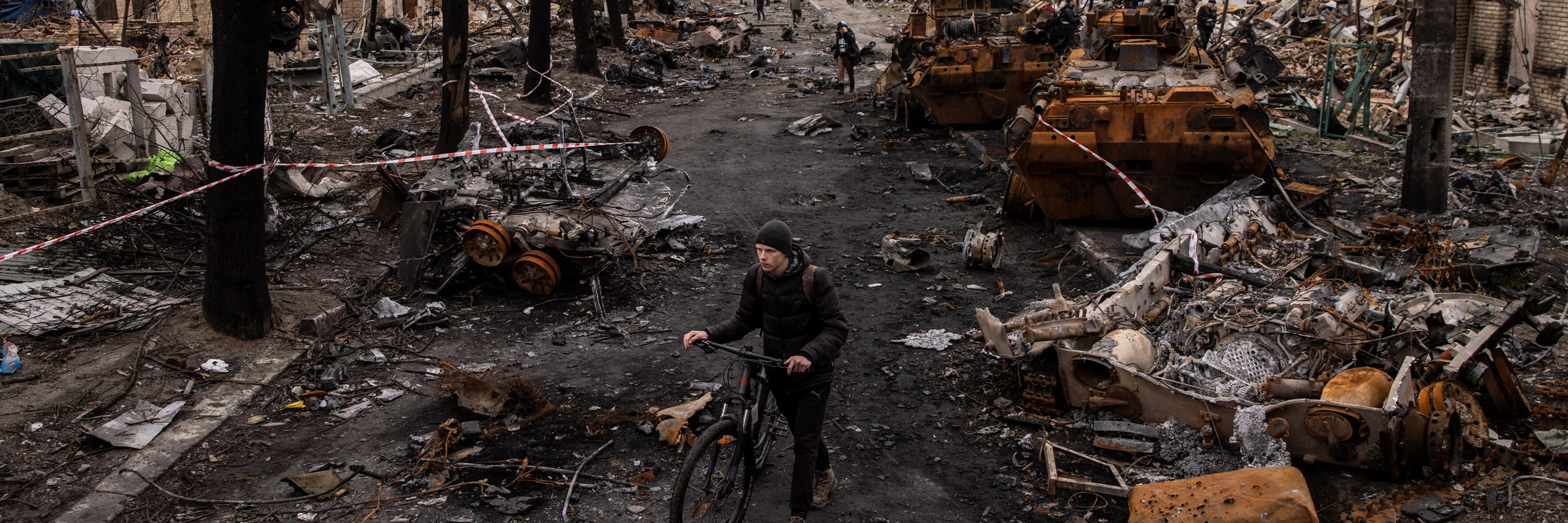 Ein Mann schiebt sein Fahrrad durch Trümmer und zerstörte Militärfahrzeuge auf einer Straße in Bucha, Ukraine. © Chris McGrath