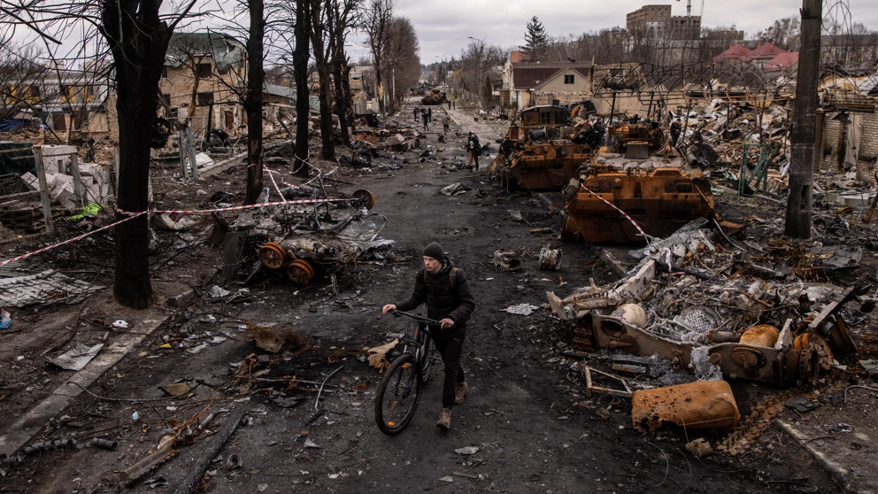 Ein Mann schiebt sein Fahrrad durch Trümmer und zerstörte Militärfahrzeuge auf einer Straße in Bucha, Ukraine. © Chris McGrath