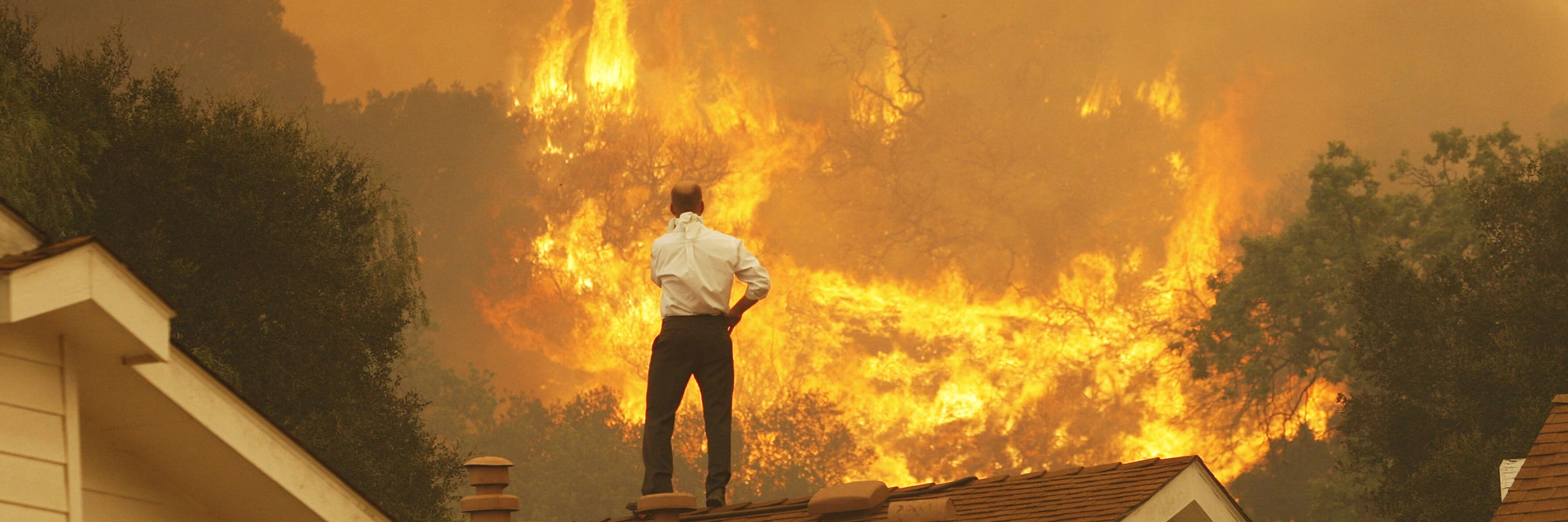 Ein Mann auf einem Dach blickt auf die nahenden Flammen eines Waldbrandes in Kalifornien – ein Bild der eskalierenden Klimakrise. © David McNew/Getty Images