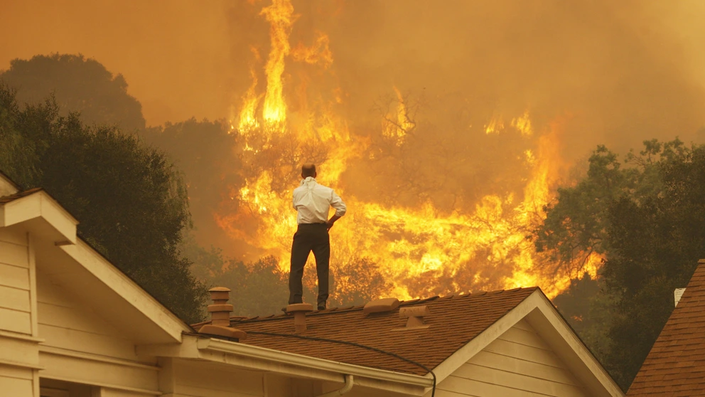 Ein Mann auf einem Dach blickt auf die nahenden Flammen eines Waldbrandes in Kalifornien – ein Bild der eskalierenden Klimakrise. © David McNew/Getty Images